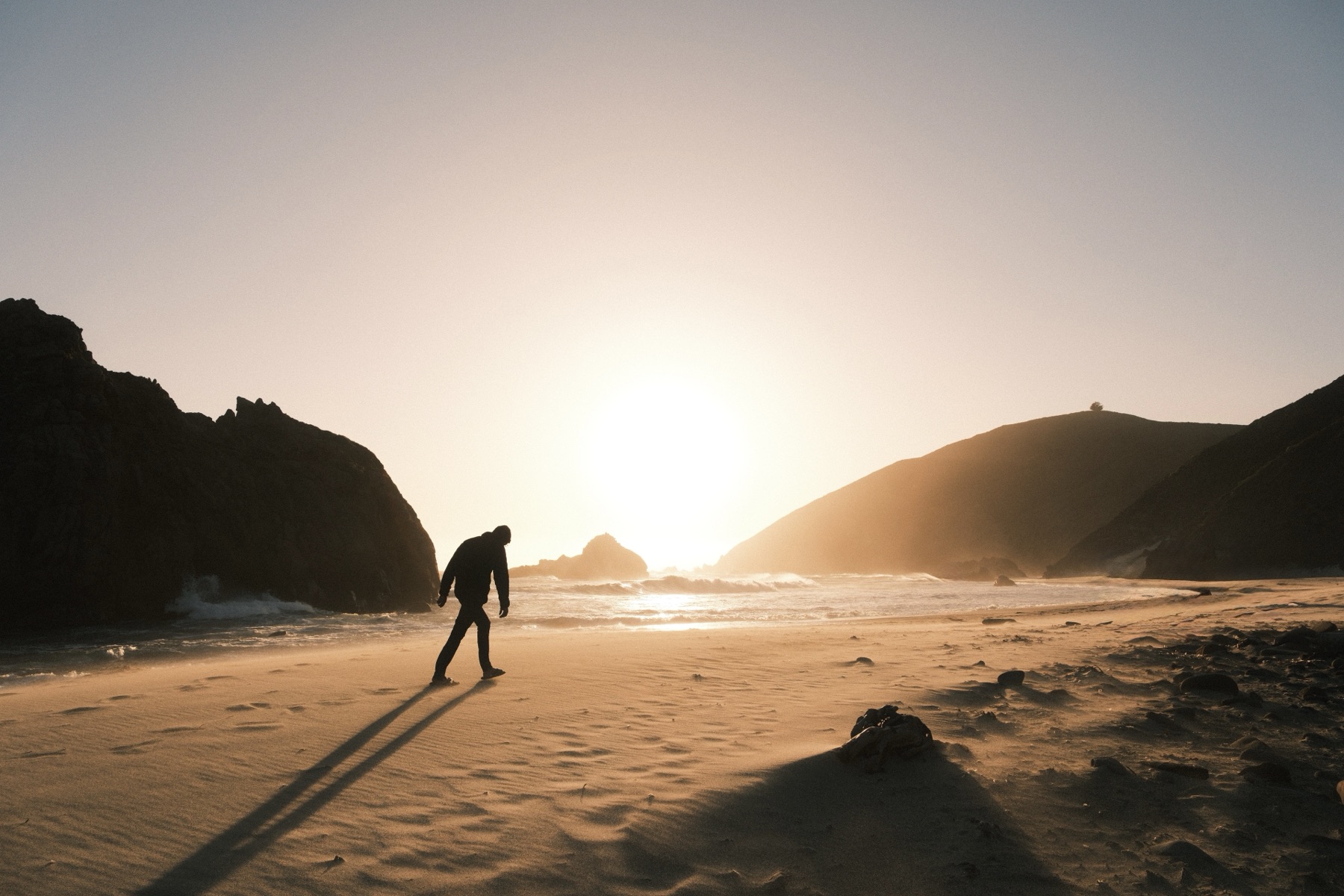 A figure walking across a sunlit beach at sunset, casting a long shadow