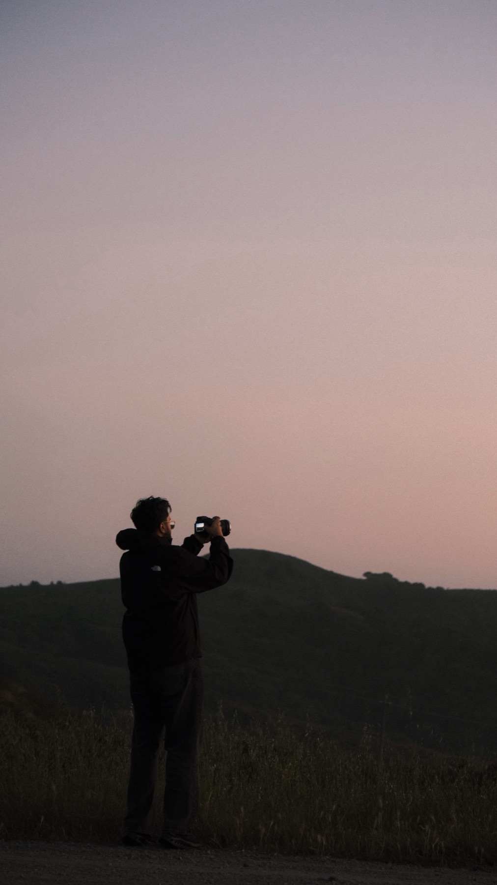 A silhouette filming with a camera on a hilltop at dusk under a pink sky